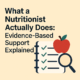 Cover image for article titled ‘What a Nutritionist Actually Does: Evidence-Based Support Explained,’ featuring a notebook with checkmarks, a magnifying glass, and an apple symbolising nutrition and assessment