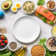 Flat lay of healthy whole foods surrounding an empty white plate, including salmon, avocado, berries, quinoa, chickpeas, oats, leafy greens, olive oil, eggs, nuts and a blue dumbbell on a light background.