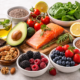 High-resolution image of fresh, nutrient-dense foods for skin health, including salmon, avocado, mixed berries, nuts, seeds, spinach, green tea, dark chocolate, and sweet potatoes arranged on a light surface.